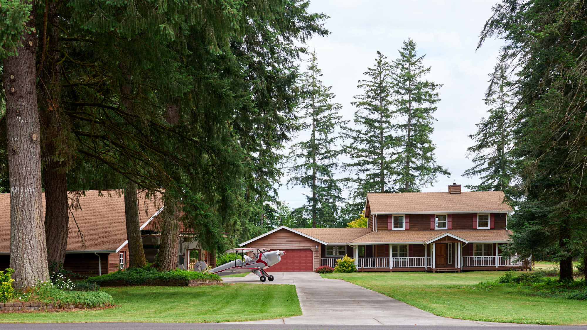 A red-and-white biplane parked at the hangar home at Parkside Airpark, cedar house and Douglas firs behind